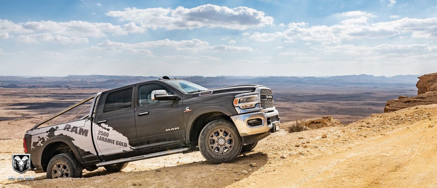 Display A white 2023 Ram 2500 Limited Longhorn Crew Cab with its headlamps on, towing a large speedboat on a dirt road.