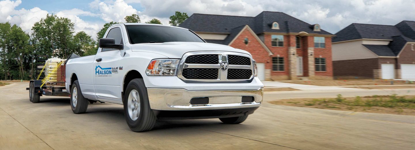 A 2022 Ram 1500 Classic with construction company signage, towing a flatbed trailer with building materials down a neighborhood street.