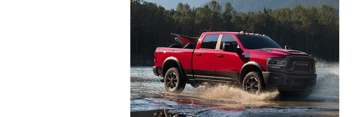 A red 2023 Ram 2500 Rebel Crew Cab carrying an ATV in its pickup box as it fords through a stream.