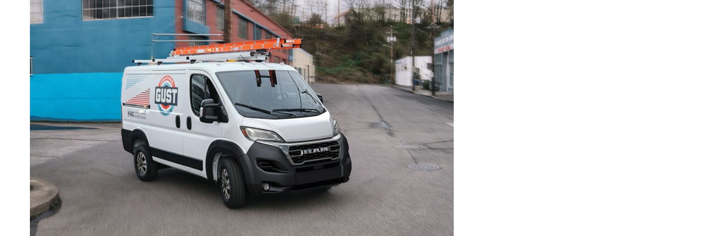 A 2023 Ram ProMaster with the logo for an HVAC company on its side and a ladder on its roof rack being driven through an industrial area.