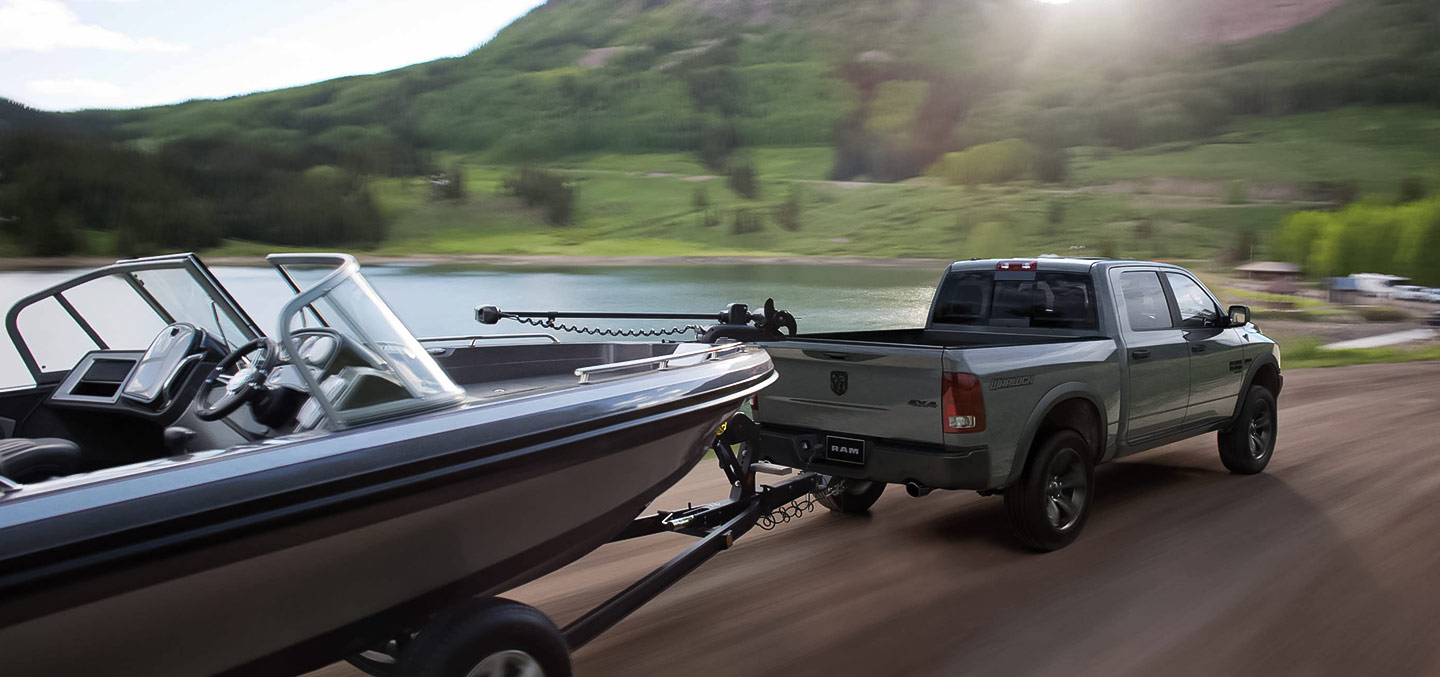Vista trasera de una Ram 1500 Classic Warlock 2023 4x4 Crew Cab circulando junto a un lago, remolcando una lancha.