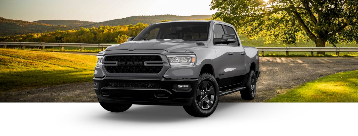 A gray 2023 Ram 1500 Big Horn Backcountry Crew Cab on a backdrop of a country road and hills of green.