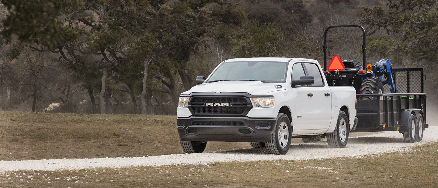 A white 2023 Ram 1500 Tradesman Crew Cab towing a tractor on a trailer.