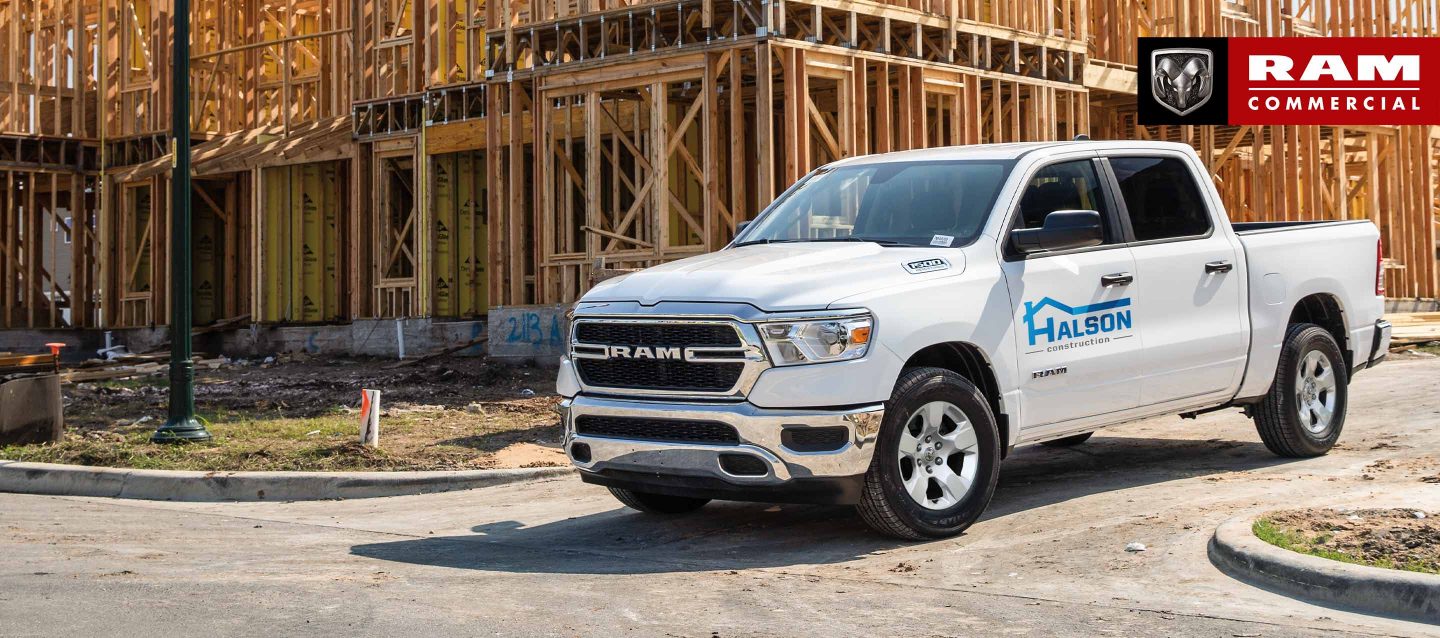 A white 2023 Ram 1500 Tradesman Crew Cab with construction company signage on its driver's door, pulling out of the driveway of a building under construction. Ram Commercial Truck Season.