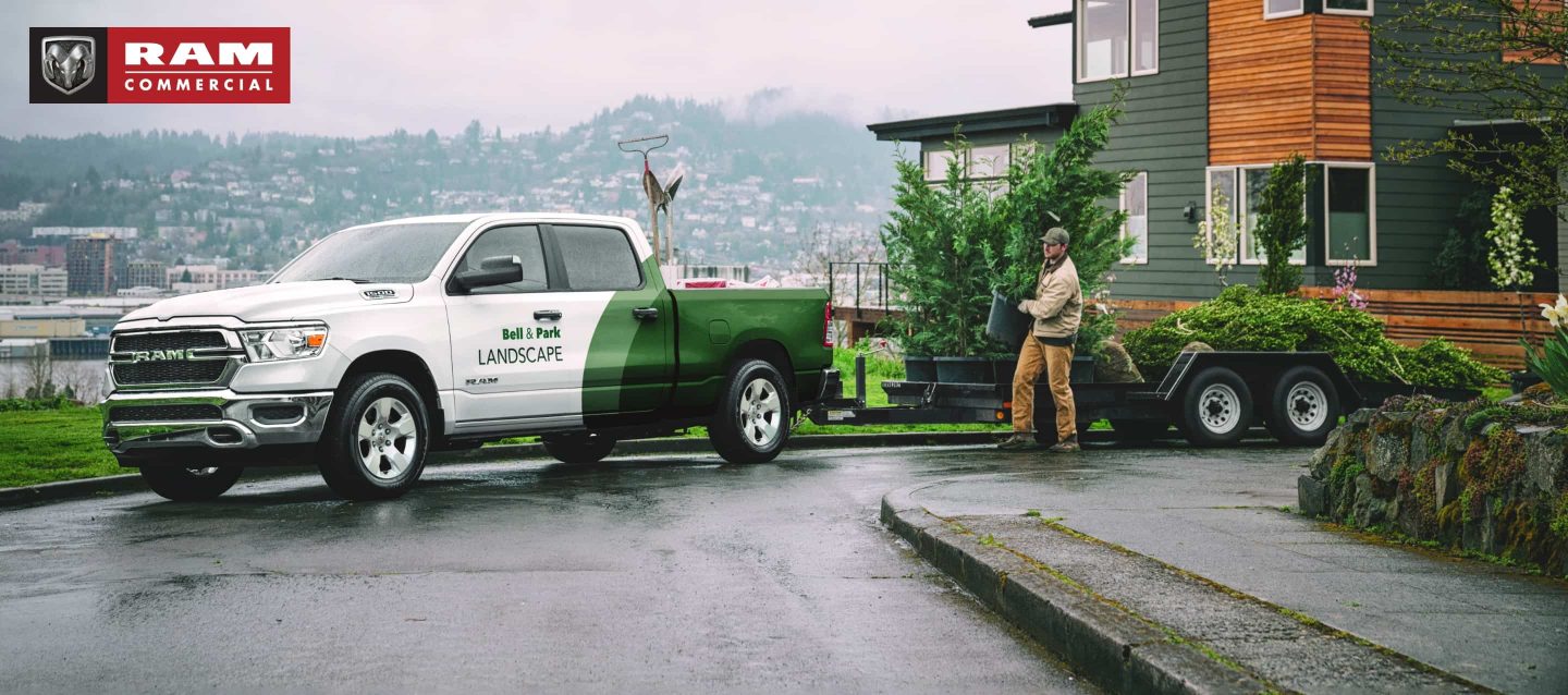 A 2023 Ram 1500 Tradesman with a landscaping business logo painted on its side parked with a trailer in front of a contemporary home. There are landscaping tools in the pickup bed and large plants on the trailer. A landscaper is pulling a potted plant off the trailer. A rainy hillside cityscape is visible in the background. Ram Commercial Truck Season.