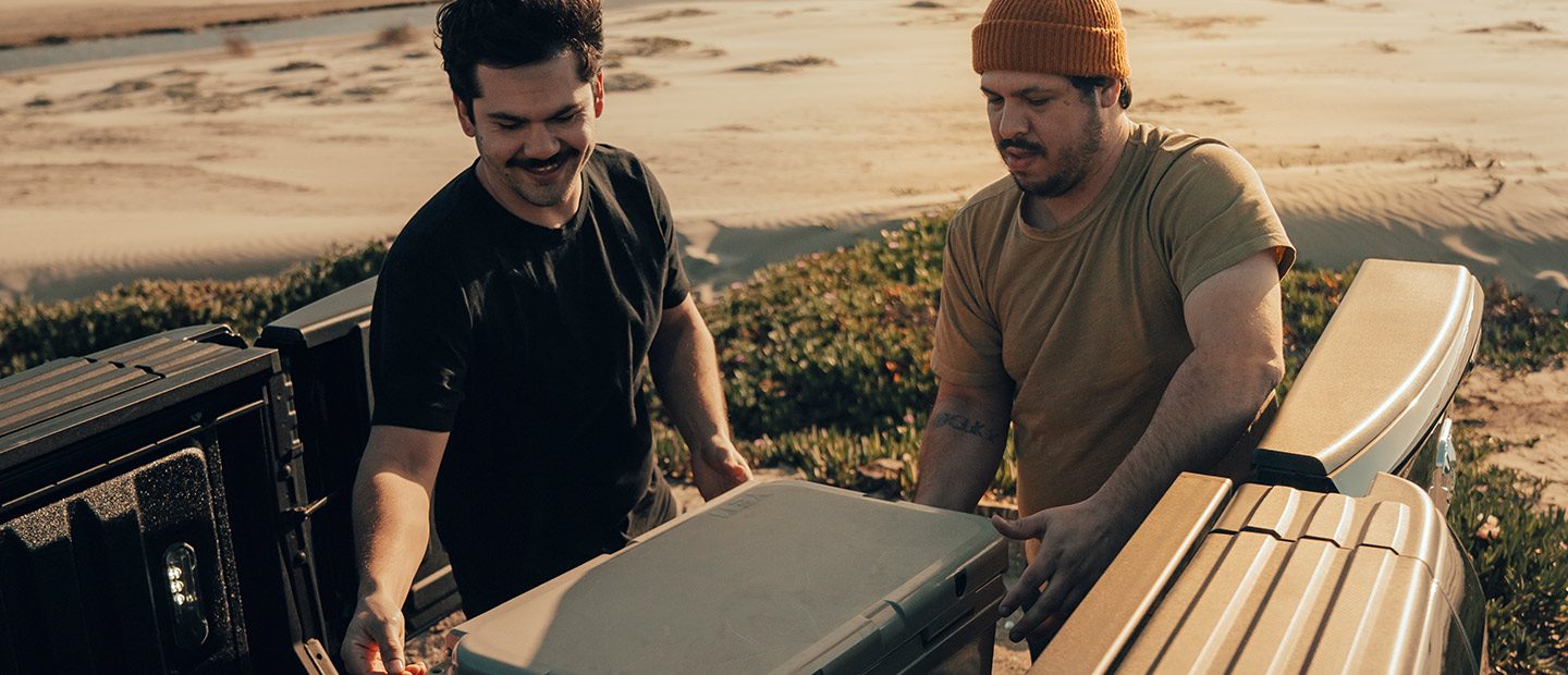 Two men loading a cooler into the bed of the 2023 Ram 1500 through the open doors of the multifunction tailgate.