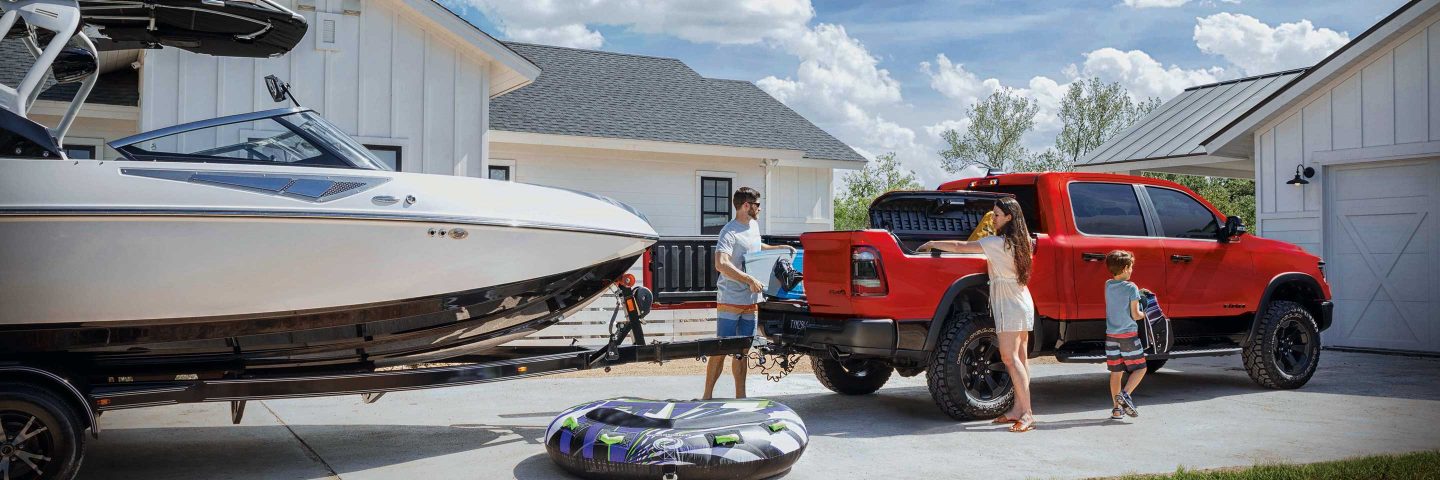 Una Ram 1500 2023 remolcando un bote de motor, y un hombre y una mujer cargando una tabla de wakeboard y flotadores en la plataforma de la camioneta y el RamBox.