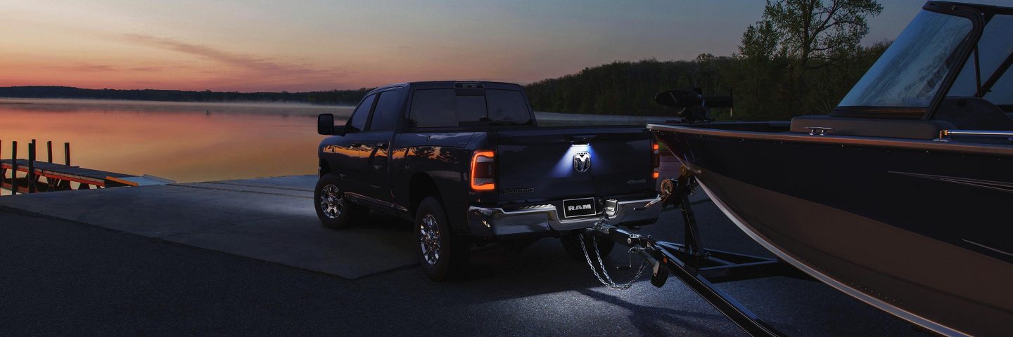 Vista trasera de una Ram 2500 Laramie 2023 con sus luces traseras encendidas, remolcando una gran lancha rápida en una rampa de botes al atardecer.