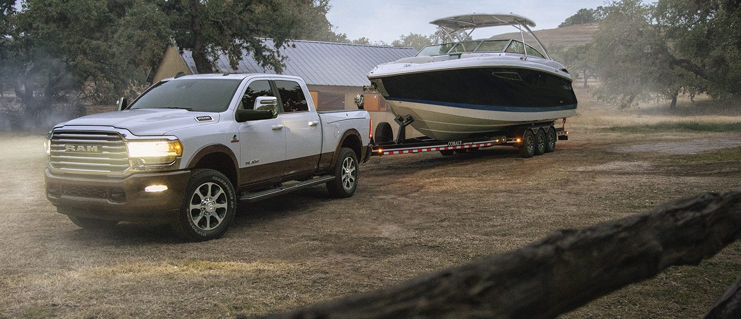 Se muestra una Ram 2500 Limited Longhorn Crew Cab 2023 blanca con los faros delanteros encendidos, remolcando una lancha grande por un camino de tierra.