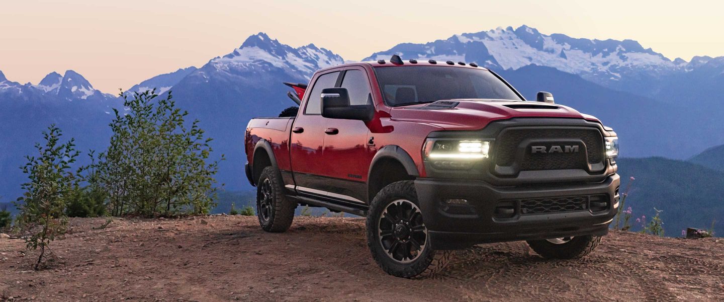 A red 2023 Ram 2500 Rebel parked on a cliff with its headlamps on and mountains in the background.