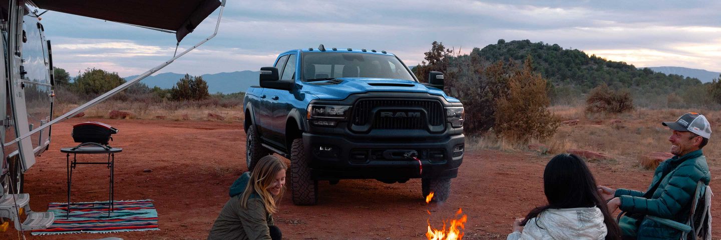 Una Ram 2500 Power Wagon 2023 azul estacionada en un campamento en el desierto al lado de una casa rodante, con tres personas sentadas alrededor de un fuego.