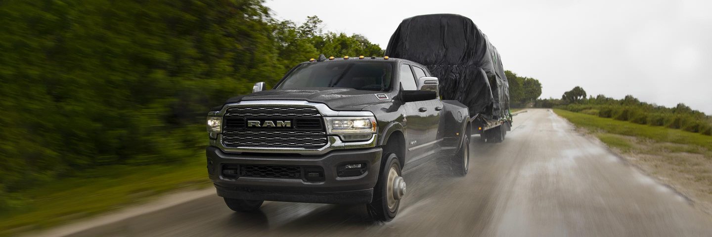 A 2023 Ram 3500 Limited Crew Cab towing a large covered trailer as it is driven down a highway in the rain.