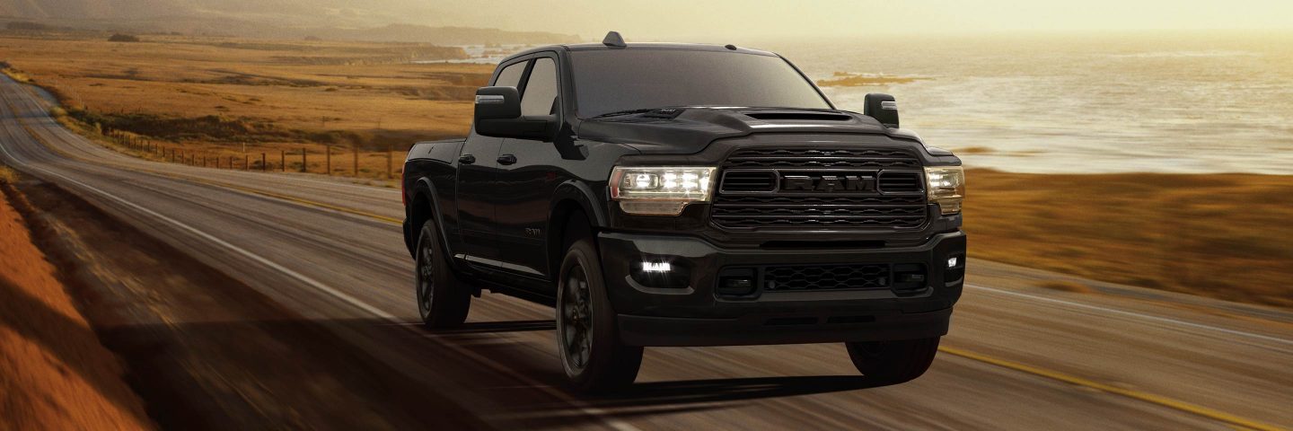 A 2023 Ram 3500 Limited Crew Cab with its headlamps on, being driven on a waterfront highway at dusk.