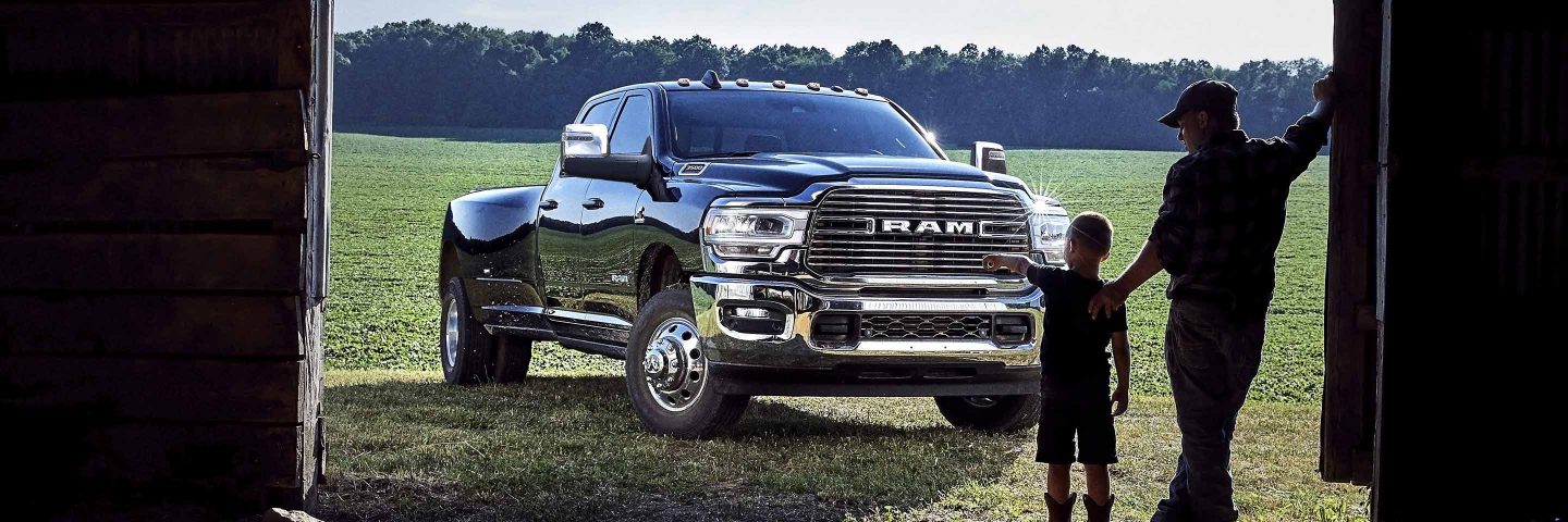 Una Ram 3500 Laramie Crew Cab 2023 estacionada en un campo de hierba junto a un granero con las puertas abiertas y un hombre y un niño parados en la entrada.