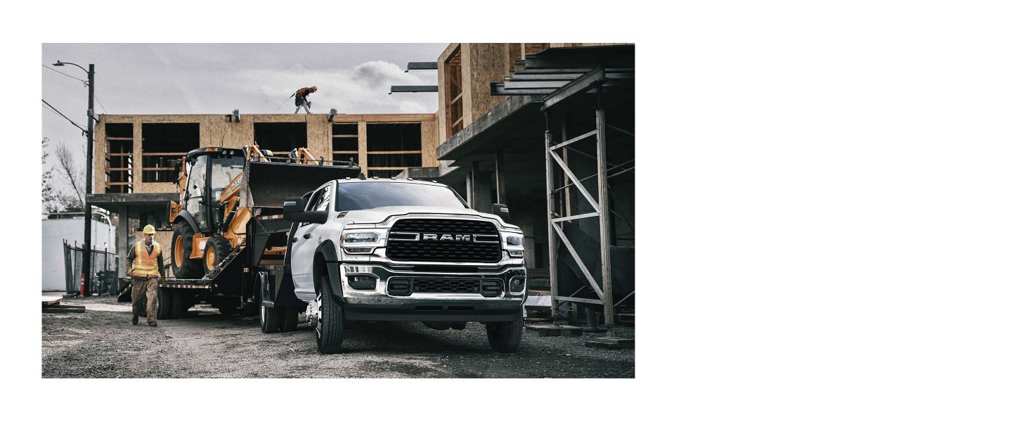 The 2023 Ram Chassis Cab towing a flatbed trailer loaded with an excavator, at a commercial construction site.