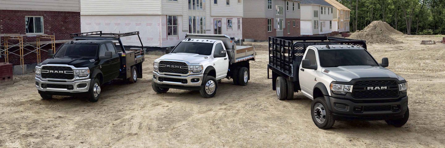 Tres Ram Chassis Cab 2023 estacionadas en una obra de construcción, cada una con una mejora distinta. De izquierda a derecha: una Ram 5500 Tradesman Chassis Cab con una mejora de plataforma con estacas, una Ram 5500 SLT Chassis Cab con una mejora de carrocería volcadora y una Ram 4500 Tradesman Chassis Cab con una mejora para paisajismo. 