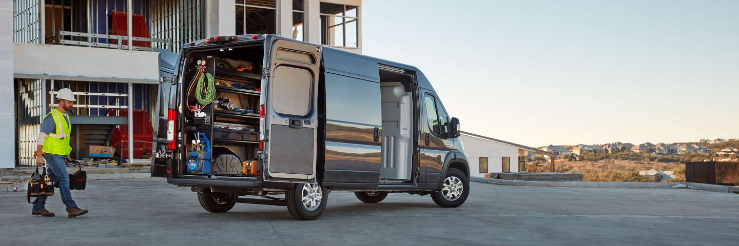 A worker carrying two toolbags approaches the open rear doors of a 2023 Ram ProMaster that has been upfitted with shelves and storage on the cargo walls.