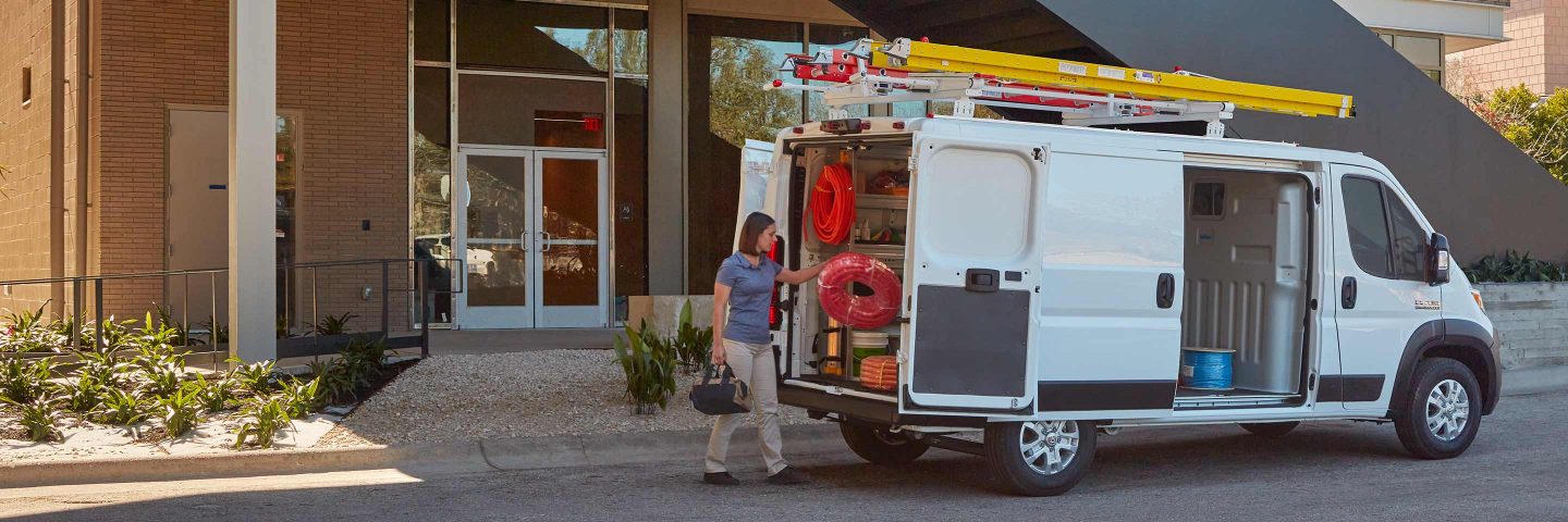 Una mujer se acerca a las puertas traseras abiertas de una Cargo Van Ram ProMaster 1500 2023 a la que se le agregaron estantes y espacio de almacenamiento como mejoras a la zona de carga.