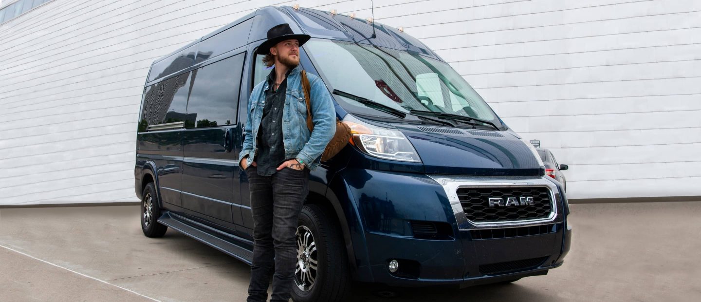 El artista country Jackson Dean de pie junto a la Ram ProMaster BandVan 2023-una Ram ProMaster 2500 High Roof Window Van con accesorios no originales.