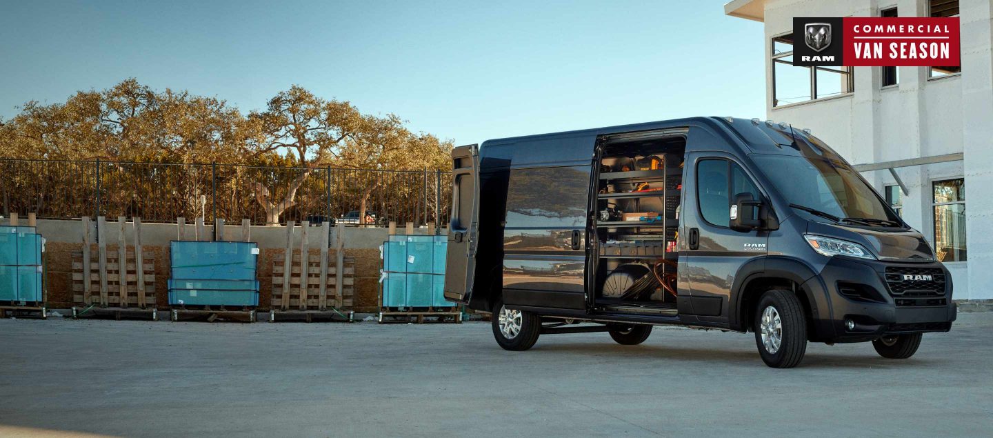 A black 2023 Ram ProMaster 3500 Cargo Van High Roof, parked beside a commercial building, with its passenger-side sliding door and rear door open, revealing shelving full of tools.