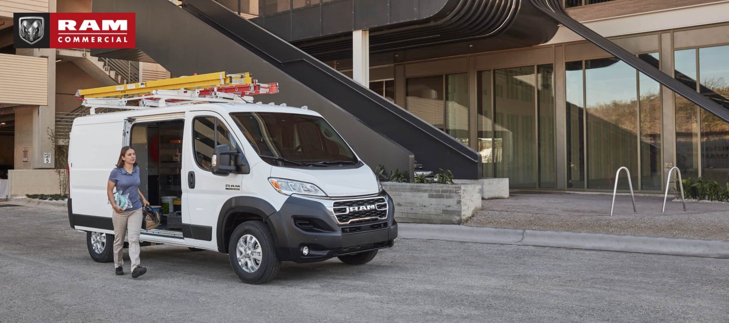 Una Cargo Van Ram ProMaster 2023 de techo bajo con escaleras sujetas a la parrilla portaequipaje, estacionada junto a un edificio comercial con la puerta lateral abierta y una mujer cargando herramientas fuera del vehículo. Temporada de vans comerciales de Ram.