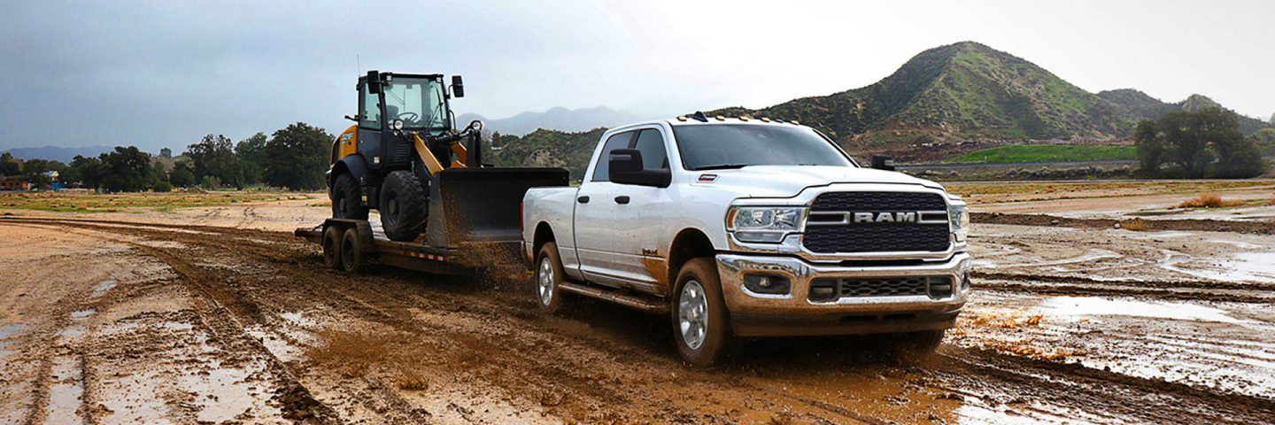 Una Ram 2500 Big Horn Crew Cab 2024 blanca remolca una pala mecánica sobre un tráiler de plataforma hacia un sitio de construcción lodoso.