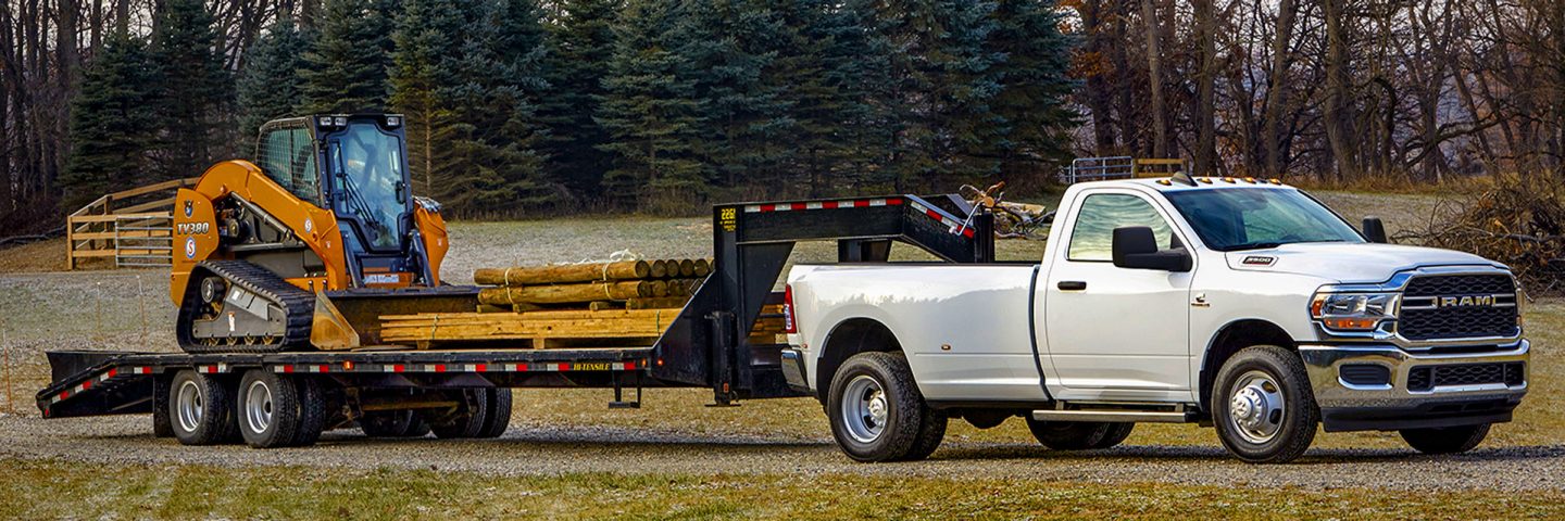 Una Ram 3500 Tradesman 4x4 Regular Cab 2024 blanca llevando un remolque de plataforma de quinta rueda cargado con una excavadora y madera.