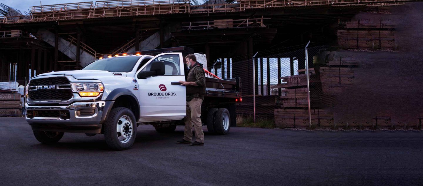 Hombre subiéndose a la cabina de una Ram 5500 Tradesman Chassis Cab Crew Cab 2024 blanca con una mejora de plataforma de estacas cargada con materiales de construcción.