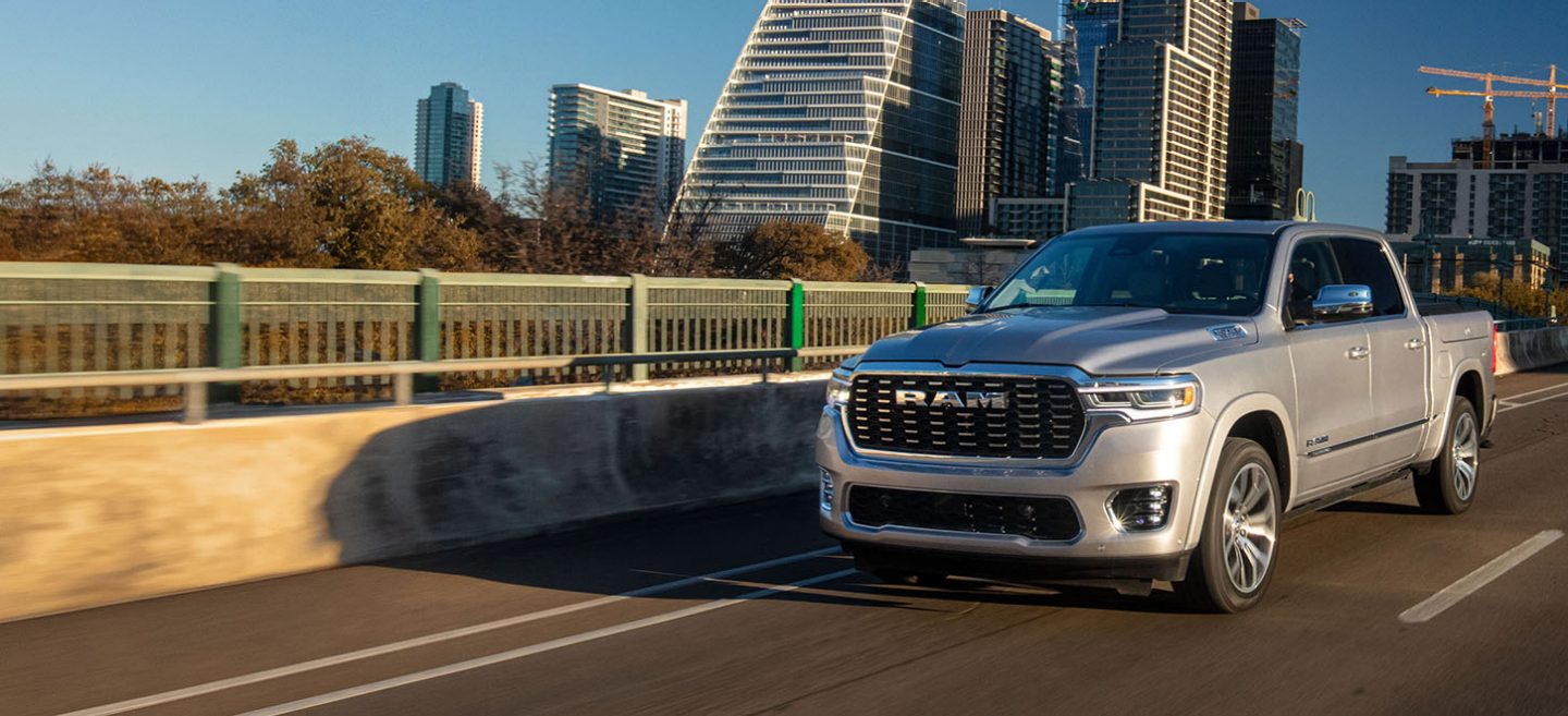 Una Ram 1500 Tungsten Crew Cab 2025 plateada circulando por un puente de una autopista en una ciudad con edificios altos de fondo.