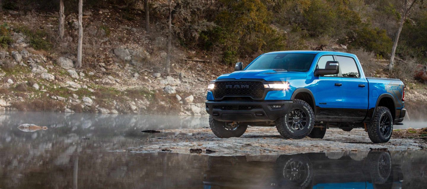 Una Ram 1500 Rebel Crew Cab 2025 azul en un camino fangoso está por entrar a un arroyo al lado de una colina grande.