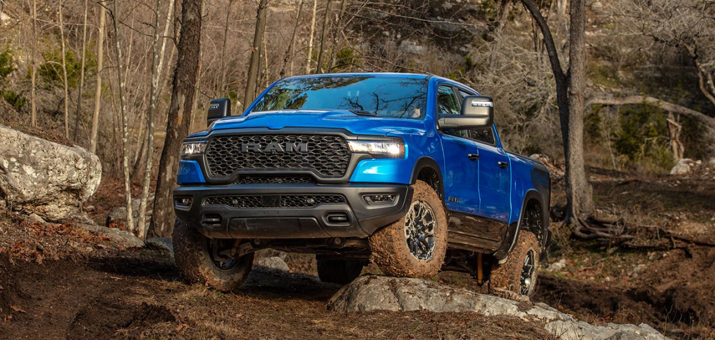 A blue 2026 Ram 1500 Rebel 4x4 Crew Cab climbing a rocky trail off-road, with a forest in the background.