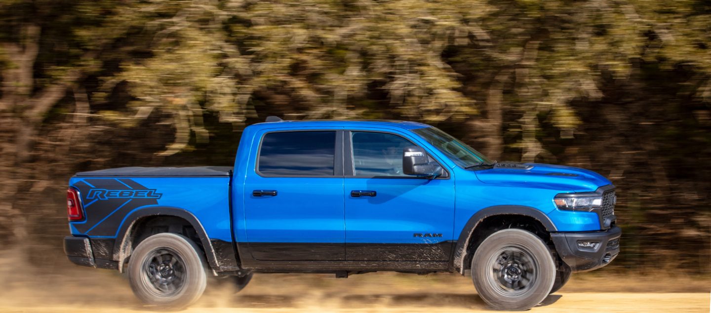 Vista de perfil del lado del pasajero de una Ram 1500 Rebel Crew Cab 2025 azul, con el fondo borroso para indicar que el vehículo está en movimiento.