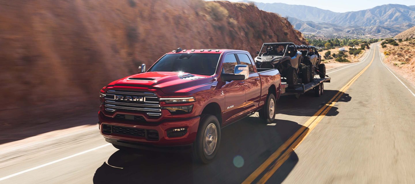 Una Ram 2500 Laramie 4x4 Crew Cab 2025 roja circulando por una carretera en las montañas, mientras remolca dos ATV.