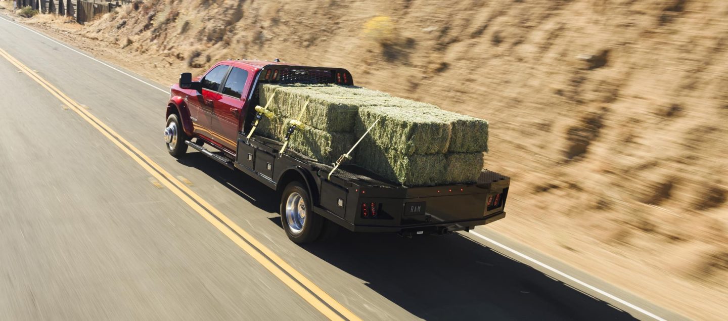 Una Ram 5500 Laramie Chassis Crew Cab 2025 roja andando por una carretera y transportando varios fardos grandes de heno sobre una plataforma larga mejorada.