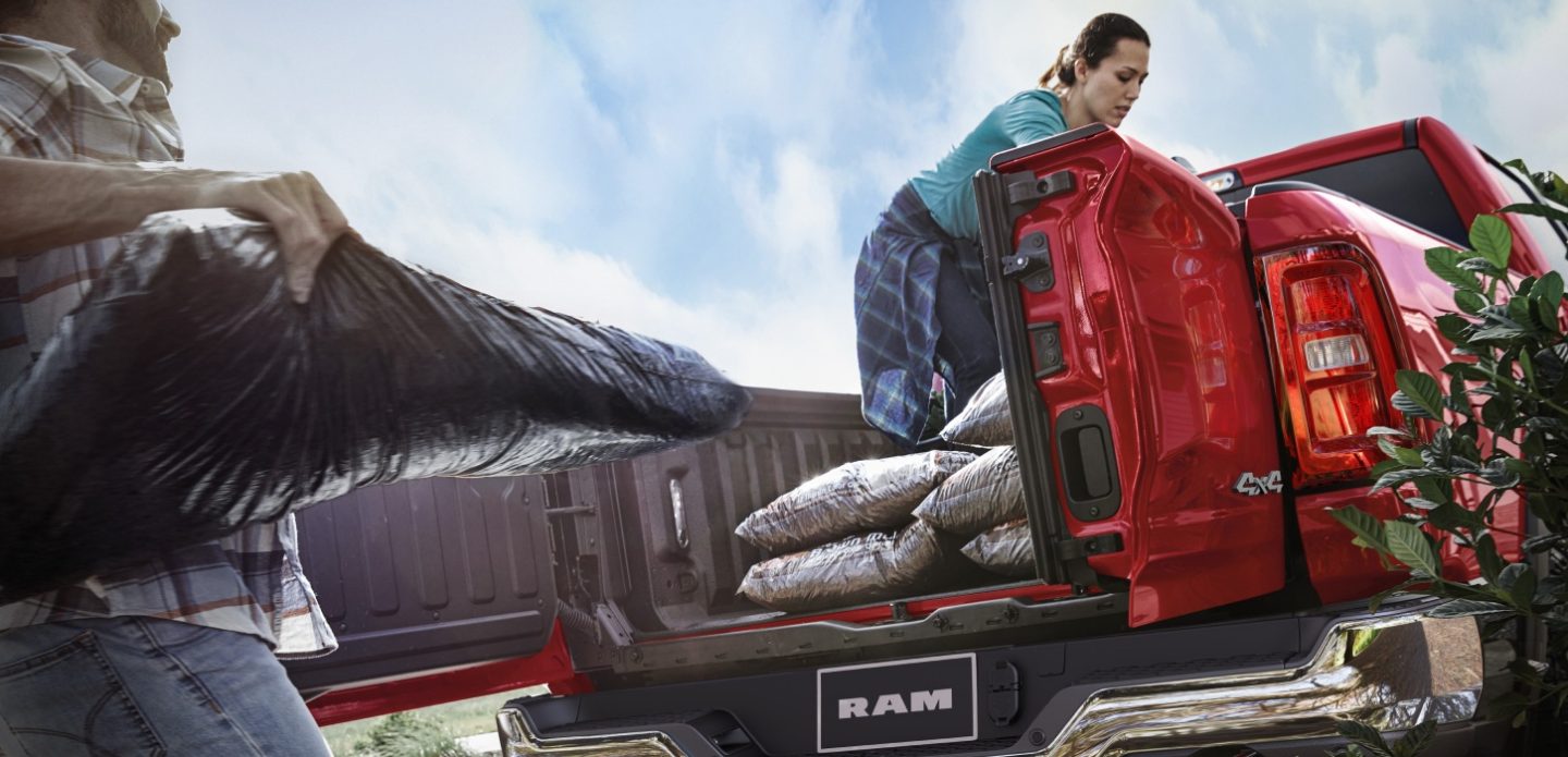 The pickup box on a red 2026 Ram 1500 Tungsten 4x4 Crew Cab with its split-door tailgate open and a woman loading bags of materials in its bed.