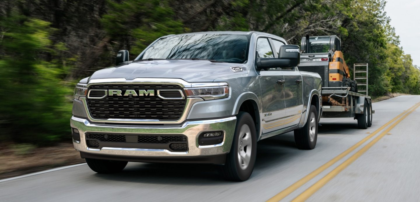 A silver 2026 Ram 1500 Big Horn 4x4 Crew Cab towing motorized equipment as it travels down a highway.