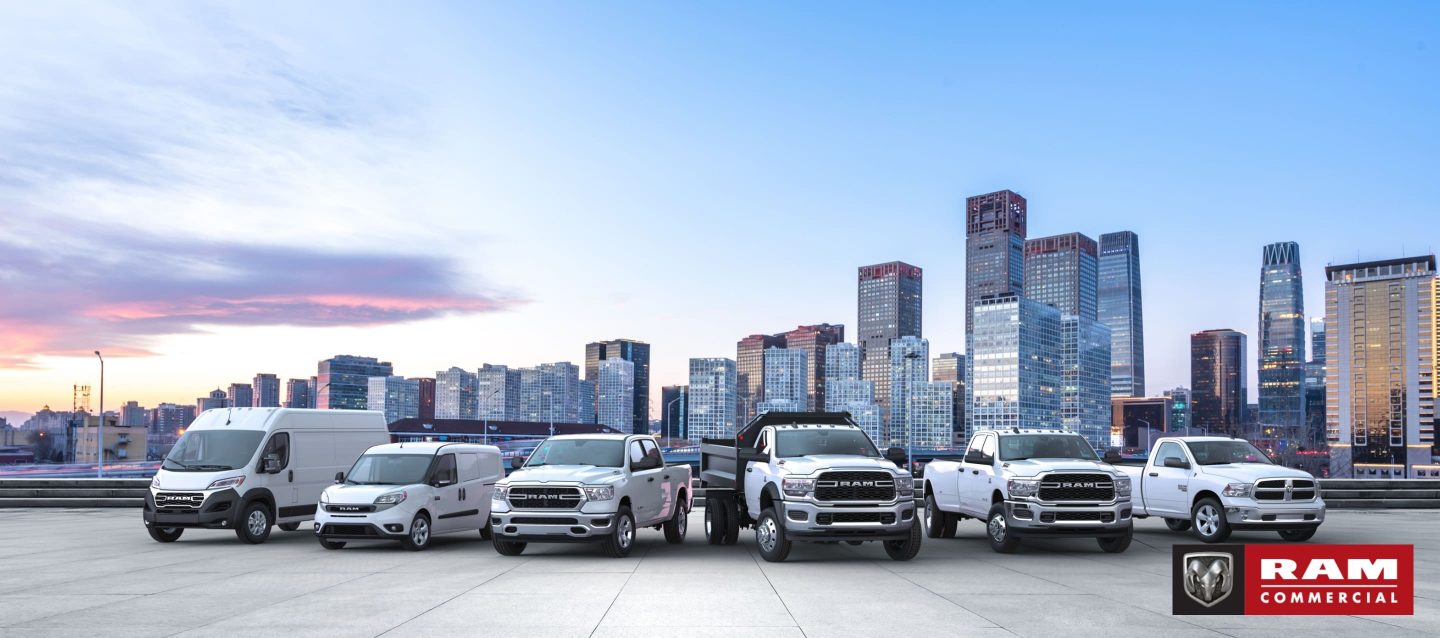 A lineup consisting of a 2023 Ram ProMaster Cargo Van, a 2022 Ram ProMaster City Passenger Van and four 2022 Ram trucks, all parked on a rooftop parking lot with a skyline of highrise buildings in the background. Ram Commercial.