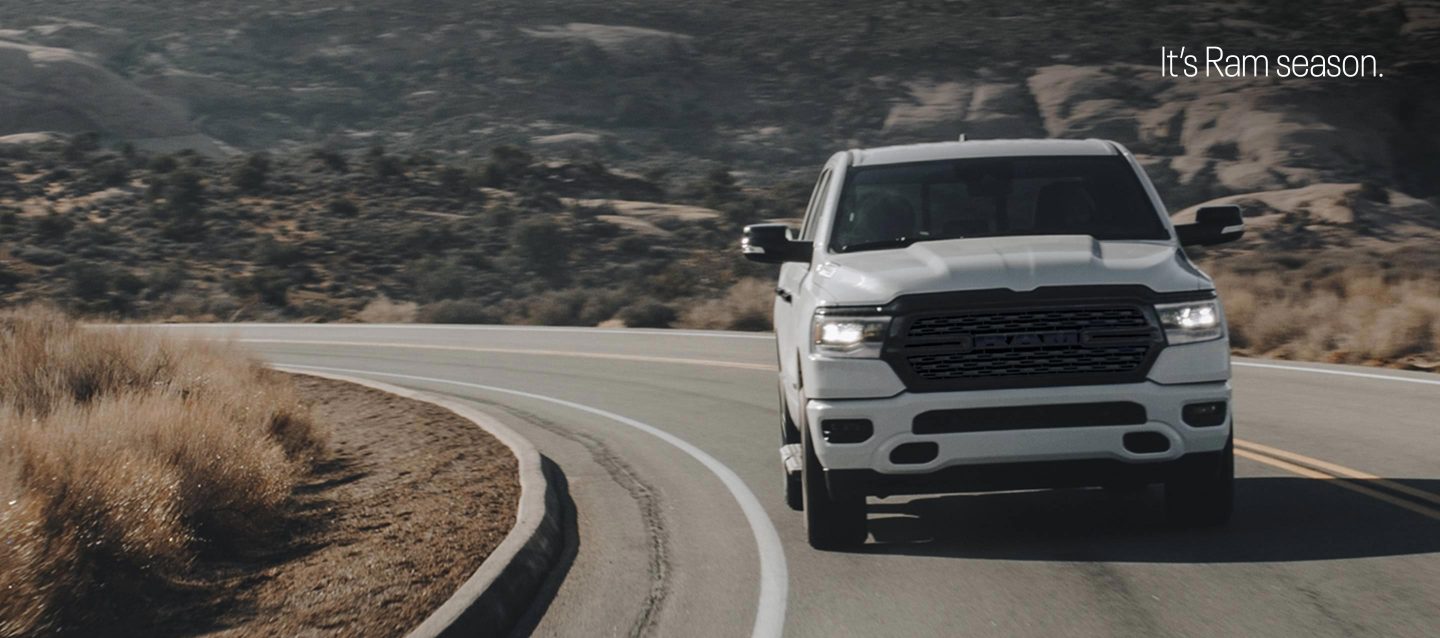 A head-on angle of a white 2023 Ram 1500 Lone Star 4x4 Crew Cab being driven up a curved mountain road in the desert. It's Ram Season.
