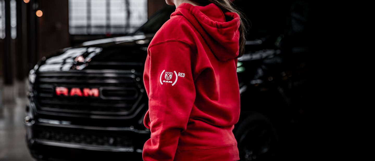 A woman wearing a red hoodie with the Ram Red logo, walking toward a 2023 Ram 1500 Limited Ram Red Edition.