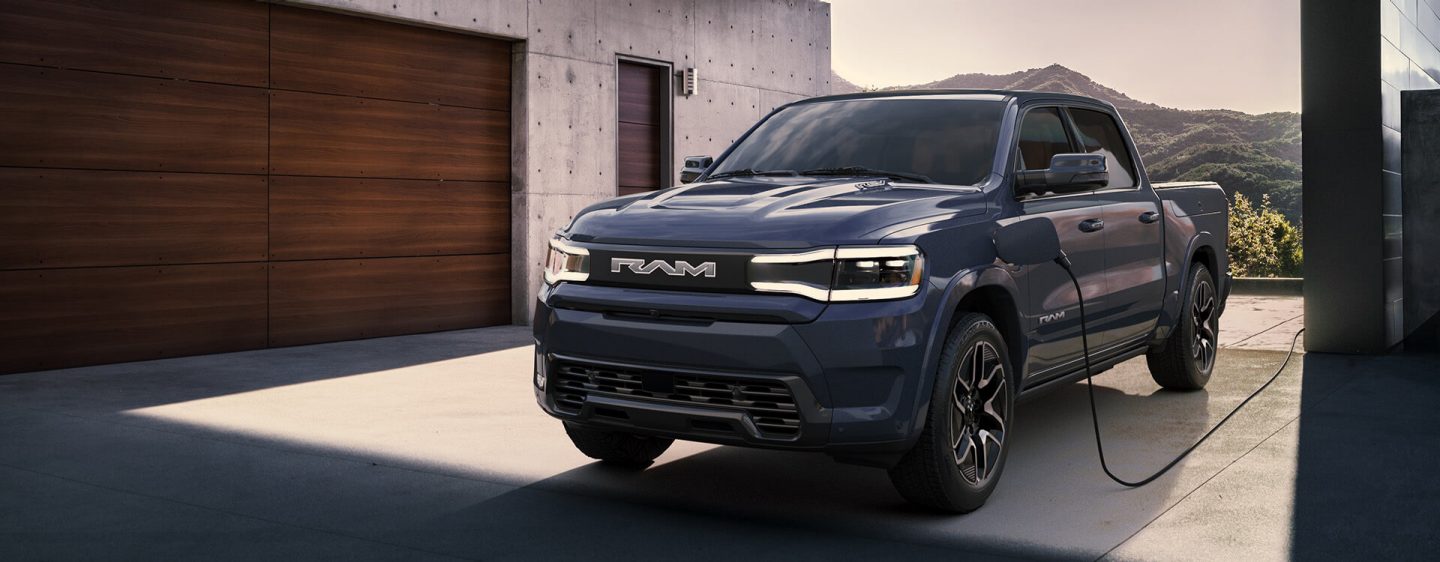 The Ram 1500 Rev Crew Cab parked in the driveway of a building with a charger cord plugged into its charging port.