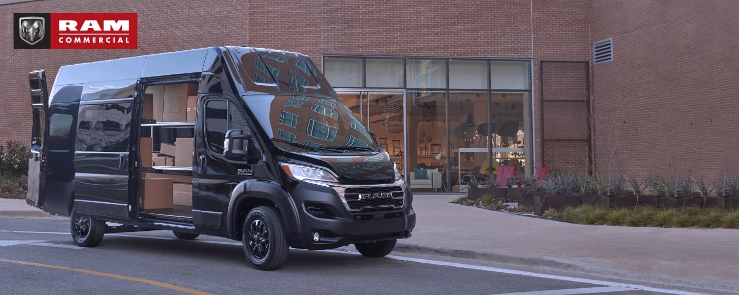 Una Ram ProMaster 3500 Super High Roof Cargo Van 2023 estacionada junto a una tienda de muebles con las puertas traseras y la puerta corrediza del lado del pasajero abiertas, dejando ver estanterías llenas de cajas. Temporada de vans comerciales de Ram.