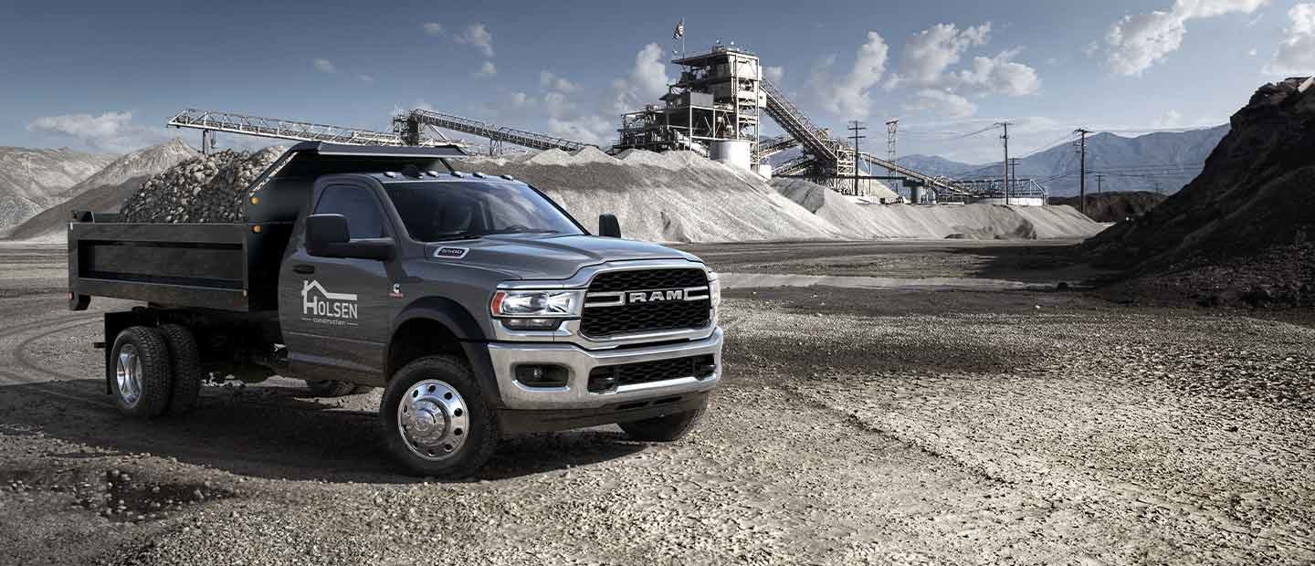 A 2023 Ram Chassis Cab Crew Cab with a gooseneck trailer filled with lumber, parked beside a forest and huge mound of cut logs, with a snow-capped mountain in the background.