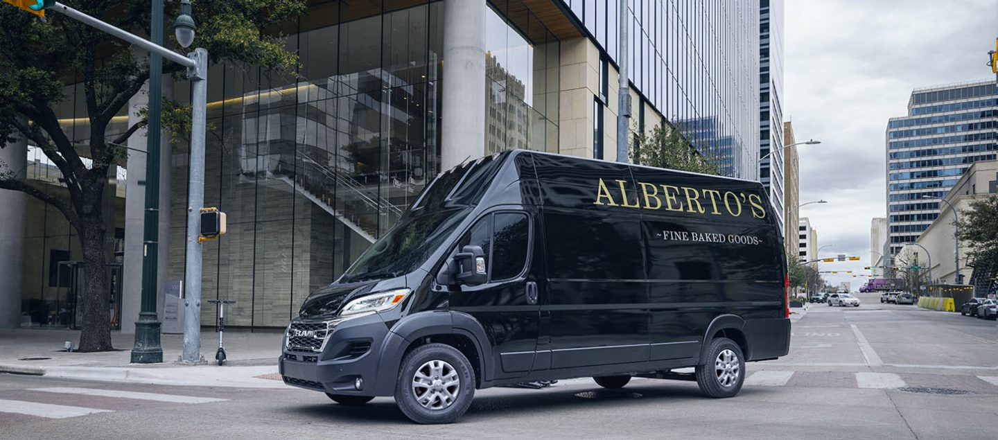 Una Ram 3500 ProMaster SLT2024 + Cargo Van Super High Roof negra con cartelería de panadería en el panel trasero del lado del conductor doblando a la derecha en una intersección con semáforo en una calle urbana.