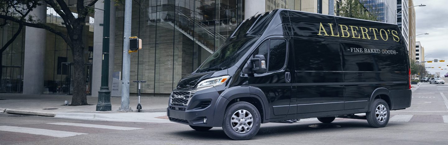 Una Ram ProMaster 2024 SLT+ Cargo Van Super High Roof negra con letreros de panadería en el panel trasero del lado del conductor, girando a la derecha en una intersección con semáforo en una calle de la ciudad.