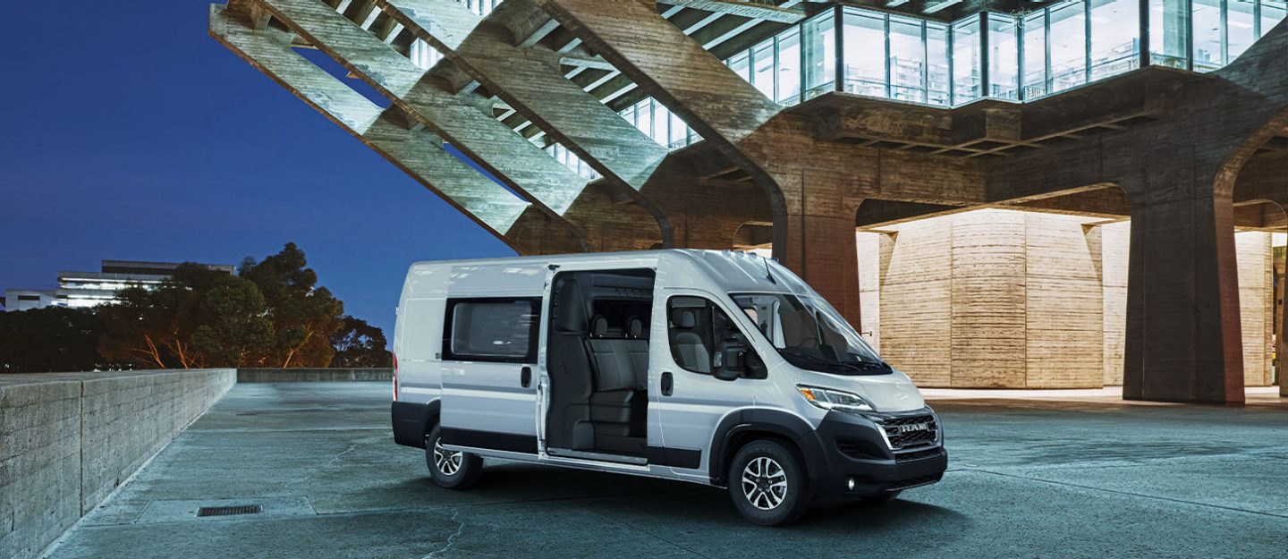 Un perfil inclinado del lado del pasajero de una furgoneta blanca Ram ProMaster 2024 2500 SLT Crew Van High Roof estacionada al lado de un edificio contemporáneo por la noche, con su puerta lateral corrediza del lado del pasajero abierta, revelando una segunda fila de asientos. 