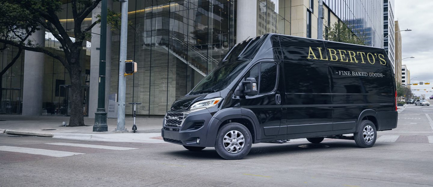 Una Ram ProMaster 3500 SLT+ Cargo Van Super High Roof 2024 negra con letreros de panadería en el panel trasero del lado del conductor, girando a la derecha en una intersección con semáforo en una calle de la ciudad.