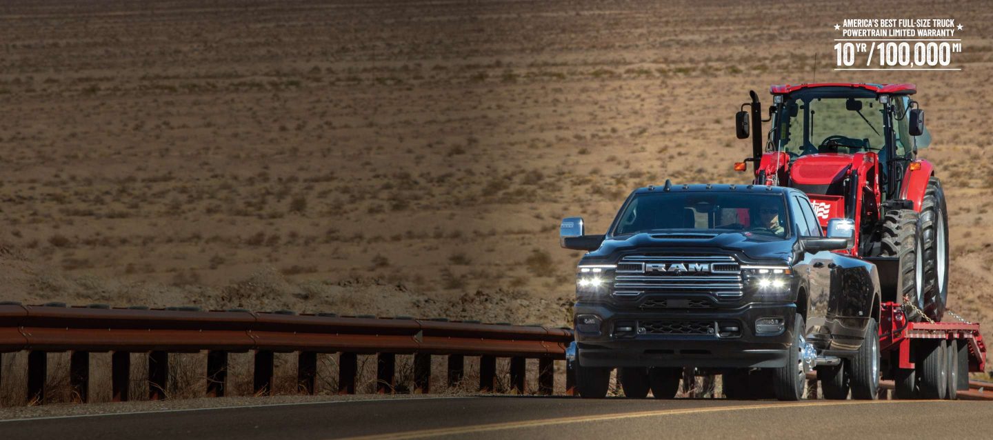 Una Ram 3500 Laramie 4x4 Crew Cab negra de 2026 remolcando un vehículo de construcción en un remolque largo mientras viaja por una carretera del desierto. La mejor garantía limitada de tren motriz de camionetas de tamaño completo de Estados Unidos: 10 años/100,000 millas.