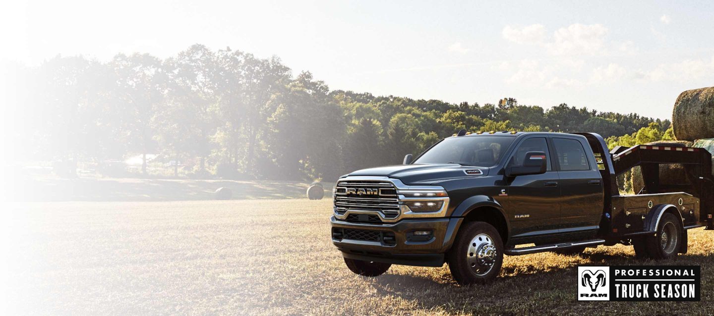 Una Ram 5500 Laramie Chassis Cab Crew Cab 2026 negra con un remolque tipo cuello de cisne cargado con varios fardos de heno, estacionada en un campo abierto. Temporada de camionetas Ram Professional.