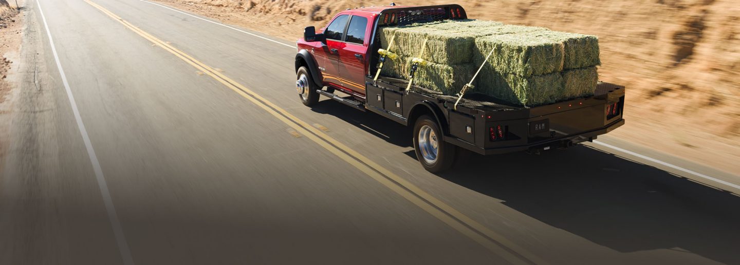 Ángulo trasero de una Ram 4500 Big Horn Chassis Cab Crew Cab 2026 roja circulando por la autopista mientras transporta varios fardos grandes de heno sobre su caja tipo plataforma larga.