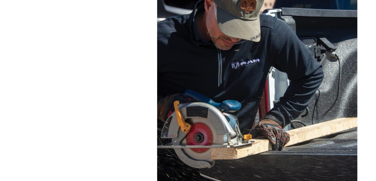 La caja de carga de una camioneta Ram 3500 2026 con un hombre usando una sierra circular conectada al inversor de la caja para cortar una tabla de madera contrachapada.